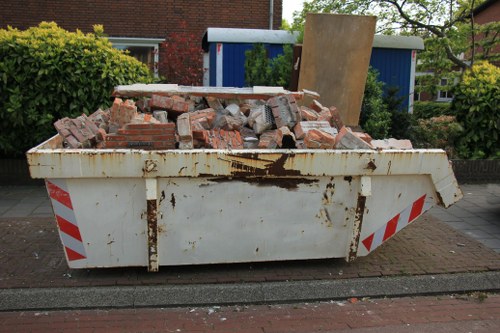 A large white industrial skip positioned on a residential street curb filled with a mixed load of red and grey bricks, some with visible mortar, and broken masonry debris. The skip shows signs of rust and dirt on its surface, with red and white reflective safety markings along its sides. It is situated on a paved driveway or road, with a background featuring residential houses, including a brick-red home partially obscured by lush green bushes, a blue shed, and a wooden panel leaning against the skip. The scene is set outdoors during daylight with natural lighting, capturing the typical environment for rubbish removal services like those offered by Office Clearance Hainault in the local area.