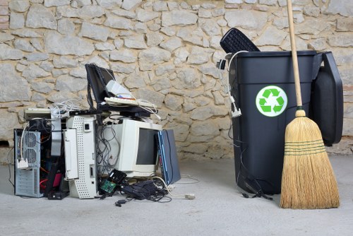 A collection of electronic waste, including old computer monitors, keyboards, and various computer parts, is stacked on the pavement in front of a stone wall; next to the pile is a large black rubbish bin with a green recycling symbol, accompanied by a wooden broom leaning against it, indicating a clean-up or rubbish removal operation close to Hainault. The scene shows various discarded devices with metal, plastic, and electronic components visible, and the outdoor setting suggests an area prepared for collection or disposal in the context of waste management services offered by Office Clearance Hainault.