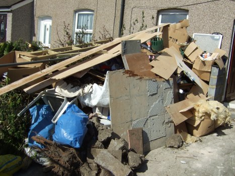 A large pile of mixed household waste and construction debris located on a pavement adjacent to a residential building in Hainault. The rubbish includes various materials such as broken concrete slabs, wooden planks, cardboard boxes, and plastic bags. Some cardboard boxes are open, revealing contents like paper and small household items, while others are flattened or partially collapsed. The concrete debris has a rough, uneven texture with exposed aggregates. Wooden planks show signs of weathering with visible grain and knots. The blue plastic bag appears full, with additional fabric or cloth materials nearby. The pile extends across the pavement, with some items leaning against the side of the building which has a light textured external wall and small windows. The environment suggests a recent or ongoing rubbish removal process from a local property, with the presence of Office Clearance Hainault providing rubbish collection services in the area.
