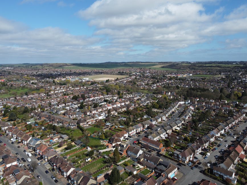 An aerial view of a suburban residential area during dusk showcases a neighborhood with closely packed houses, each with rooftops primarily in dark grey and brown finishes, surrounded by green trees a