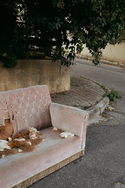 A worn and dilapidated light blue upholstered sofa with torn fabric, exposed foam, and sagging cushions placed on a concrete pavement against a graffiti-marked brick wall. The sofa's armrests and back
